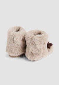 Dunstiga beige tofflor med en mjuk textur, ankellängd och en brun pom-pom-accent på sidan. Platt sula för komfort på inomhusytor.