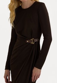 Tmavě hnědé přiléhavé šaty s dlouhými rukávy a drapovaným designem, které jsou doplněny hnědým koženým páskem se zlatou sponou.