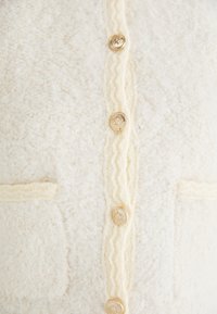 Krämfärgad cardigan gjord av textil med struktur. Den har en knäppning framtill med gyllene knappar och två påsydda fickor. Zickzackdetalj som accent.