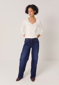 Frau mit lockigem Haar, die mit den Händen hinter dem Rücken steht, trägt ein weißes langärmeliges Oberteil, dunkelblaue weite Jeans und braune spitze Schuhe.