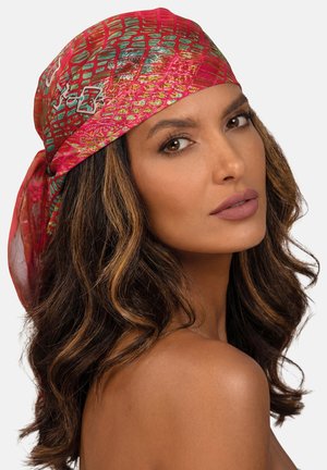 Kobieta z falującymi brązowymi włosami, ubrana w czerwony apaszka we wzory, spogląda przez ramię z neutralnym wyrazem twarzy na jednolitym tle.