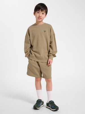 Junge steht mit den Händen in den Taschen, trägt einen beige übergroßen Sweatshirt und Shorts, weiße Socken und dunkelgrüne Sneakers vor weißem Hintergrund.