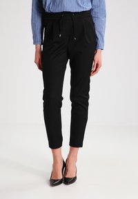 Pantaloni neri affusolati con vita elasticizzata e coulisse, abbinati a una maglietta a righe blu. Il tessuto sembra morbido ed elasticizzato.