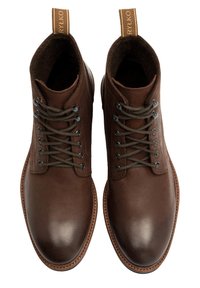 Brązowe skórzane botki do kostki z okrągłym noskiem, sznurowanym fasonem i kontrastowymi brązowymi sznurowadłami. Posiadają fakturowany kołnierz i pętlę do zakupu.