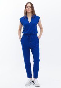 Blå todelt outfit med en kortærmet top med V-hals og bukser med snøre. Stoffet ser tekstureret og tætsiddende ud.