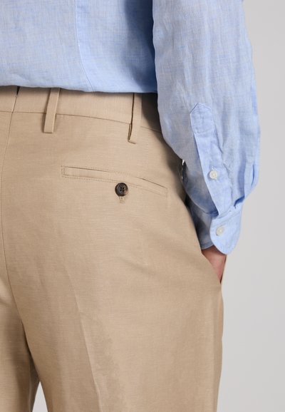 Gros plan d'une personne portant un pantalon beige avec une poche arrière boutonnée et une chemise à manches longues bleu clair rentrée.