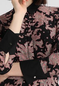 Svart blus med ett livligt rosa paisleymönster, med en strukturerad krage och kontrasterande svarta manschetter, accentuerad med runda knappar.