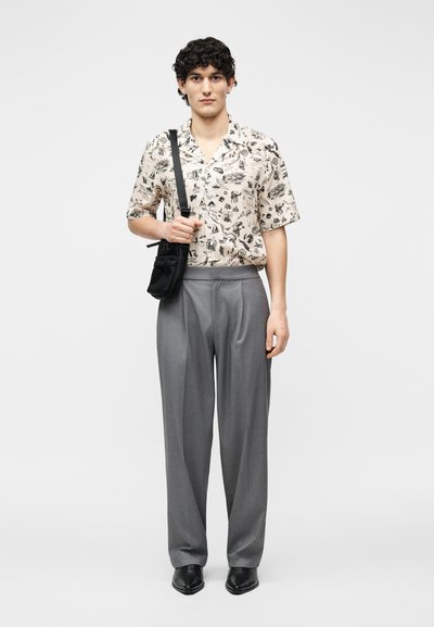 Νέος άνδρας με πουκάμισο με σχέδιο και κοντά μανίκια, γκρι παντελόνι, μαύρα παπούτσια, κρατώντας μαύρη τσάντα χιαστί με λουρί στον αριστερό ώμο.