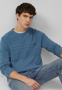 Pull bleu avec des rayures horizontales texturées, col rond, tissu en maille, et petite étiquette de marque sur la poitrine. Porté avec un jean en denim clair.