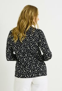 Camisa negra con un patrón moteado en blanco y dorado, de mangas largas, fabricada en un tejido suave, de corte relajado y con dobladillo recto. Lleva puesta sobre pantalones blancos.