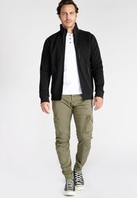 Zwarte fleece jack met ritssluiting en opstaande kraag, witte shirt eronder, olijfgroene cargobroek met ritszakken, en zwarte sneakers.