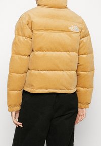 Beige kordflöjls puffer-jacka med The North Face-logotyp på baksidan, bärs av en person i svarta byxor, visad bakifrån.