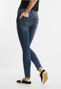Vaqueros ajustados en denim azul oscuro con una textura desgastada, que incluyen bolsillos traseros y costuras visibles, combinados con zapatillas negras.