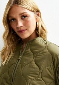 Chaqueta bomber acolchada en verde oliva con cuello alto, cierre de cremallera en la parte delantera y patrón de ondas texturizado. Tejido suave con un acabado liso.