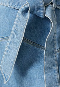 Vaaleansininen farkkukangas, jossa solmittu solmu. Sisältää hienovaraisia tekstuureja ja ompeluaksentteja, korostaen kankaan pehmeyttä ja rakennetta.