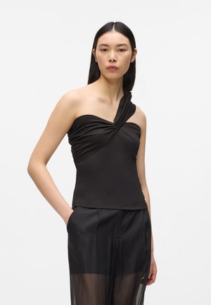 Femme portant un haut noir asymétrique à bretelle torsadée et un pantalon noir transparent, debout avec une main dans la poche sur fond blanc.