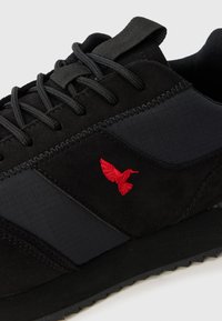 Black athletic shoe with a textured suede and smooth fabric upper, featuring red embroidered logo, black laces, and rubber outsole.