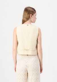 Beige sleeveless top with a square cut and back closure. Features a decorative bow detail and subtle texture. Paired with light patterned pants.