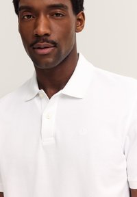 Nahaufnahme eines Mannes, der ein weißes Polohemd mit zwei Knöpfen und einem dezenten, gestickten Logo auf der Brust trägt, vor einem neutralen Hintergrund.