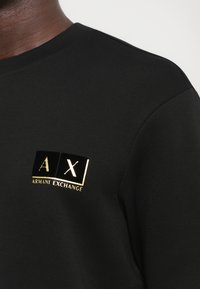 Close-up of black Armani Exchange shirt featuring gold "A X" logo and "Armani Exchange" text on the chest.