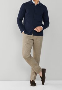 Cardigan blu navy con bottoni, colletto di camicia azzurro chiaro visibile sotto, abbinato a pantaloni di lino beige e mocassini marrone scuro.