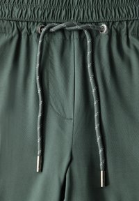 Green fabric shorts with an elastic waistband, featuring a drawstring with metallic tips and a side pocket. Smooth texture, casual design.