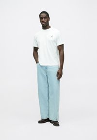 Biała koszulka z logo, zestawiona z szerokimi, jasnoniebieskimi dżinsami. Modelka ma na sobie czarne obuwie, stoi na jednolitym tle.