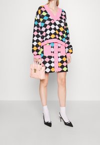 Kleurvolle gebreide trui met zwarte, roze en meerkleurige ruitpatronen, gecombineerd met een bijpassende rok met roze knopen en zakken, en zwarte puntige hakken.