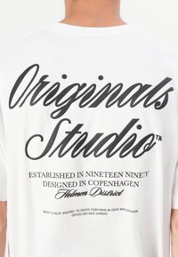 Bílý bavlněný trik s velkým černým grafickým nápisem na zadní straně, včetně textu "Originals Studio" a dalších designových detailů níže.