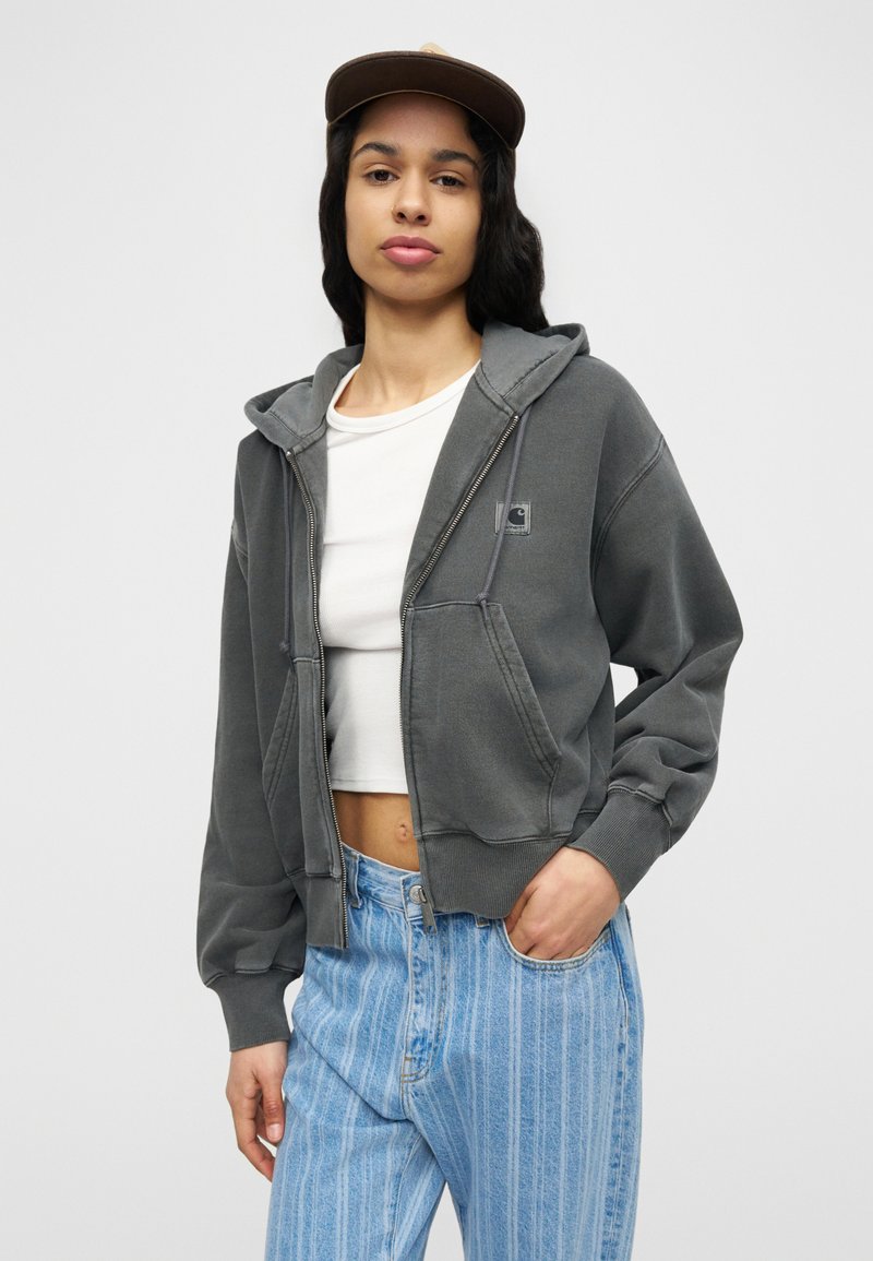 Giovane con cappellino marrone, felpa grigia con zip, top bianco corto e jeans a righe blu a vita alta, in piedi con una mano in tasca.