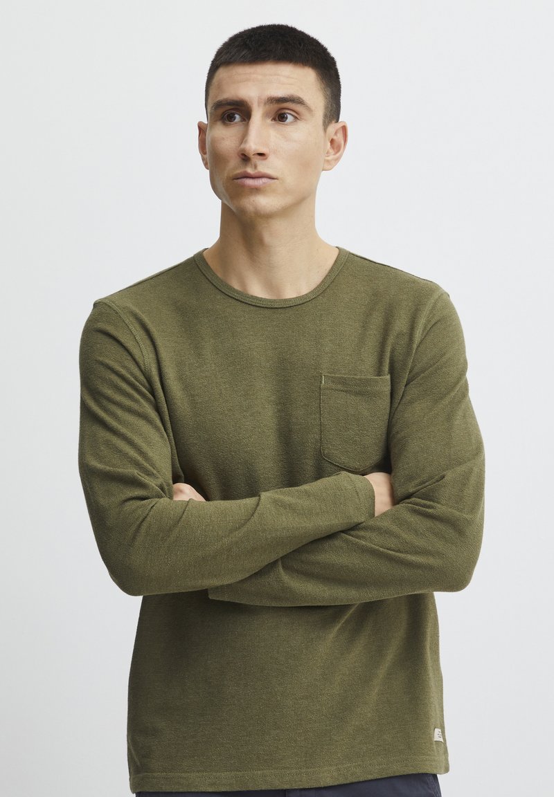 Olive-green long-sleeve t-shirt in a textured fabric, featuring a crew neckline and a chest pocket. Simple, fitted design.