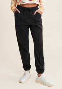 Pantalones joggers negros ajustados con cintura elástica, bolsillos laterales y puños acanalados, combinados con zapatillas blancas. El material parece suave.