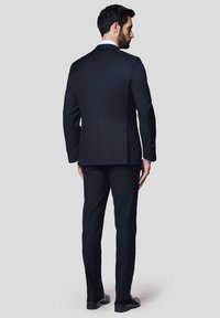 Ciemnogranatowy garnitur o dopasowanym kroju, z pojedynczym rozcięciem i kontrastową białą koszulą; w połączeniu z czarnymi, wypolerowanymi butami. Widok z tyłu.