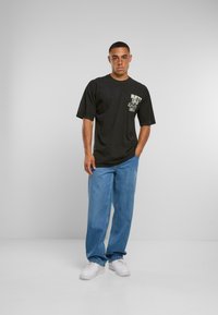 Černé tričko s krátkým rukávem a grafickým potiskem na hrudi, v kombinaci s volnými modrými džínami a bílými teniskami. Jednoduchý design a střih.