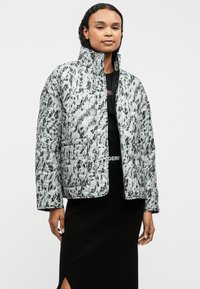 Quilted puffer jacket σε ανοιχτό γκρί χρώμα με μαύρα αβέβαια σχέδια, ψηλό γιακά, μπροστινό φερμουάρ και πλευρική τσέπη, φορεμένο πάνω από ένα μαύρο τοπ και φούστα.