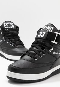 Baskets montantes noires avec semelles blanches, "EWING 33" sur les lanières et la signature "Patrick Ewing" sur les côtés.