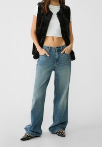 Zwart nepbontvest over een wit cropped shirt, gecombineerd met losse blauwe spijkerbroeken en zwarte puntschoenen met studs.