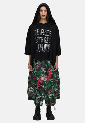 Femme portant une veste à capuche noire par-dessus un t-shirt noir avec un motif graphique, un pantalon cargo ample à motif camouflage floral vert, rouge et gris, et des bottes noires.