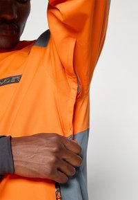 Άνδρας που φοράει ένα πορτοκαλί και γκρι μπουφάν, ανοίγει έναν φερμουάρ αερισμού κάτω από το σηκωμένο χέρι του, μπροστά σε ένα μονόχρωμο φόντο.