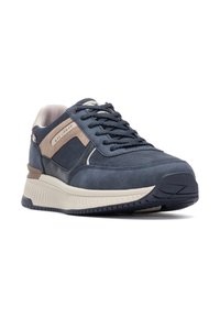 Scarpa sportiva blu con texture scamosciata, dotata di accenti beige, design con allacciatura, e una spessa suola bianca con un motivo di grip testurizzato.