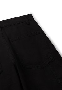 Acercamiento de un bolsillo de pantalones negros con costuras visibles y un pequeño logotipo bordado cerca de la esquina inferior.