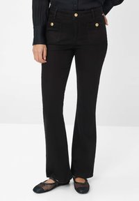 Schwarze, ausgestellte Jeans mit zwei vorderen Taschen, goldfarbenen Knöpfen und dezentem Steppdetails. Das Material ist elastischer Denim. Flache Schuhe sind sichtbar.