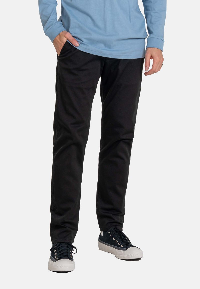 Hombre con pantalones negros, zapatillas blancas y negras, y camisa de manga larga azul, con una mano en el bolsillo sobre fondo blanco.