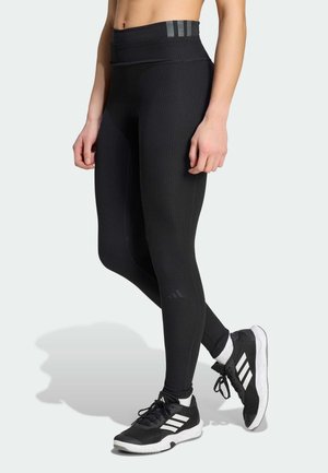 Žena v černých žebrovaných legínách a černých teniskách Adidas s bílými pruhy stojí se zkříženýma nohama na světle pozadí.