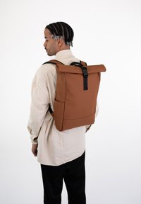 Rucsac brun cu capac rulabil, dotat cu catarame și curele negre, având o textură netedă și un design minimal, purtat pe spatele unei persoane.