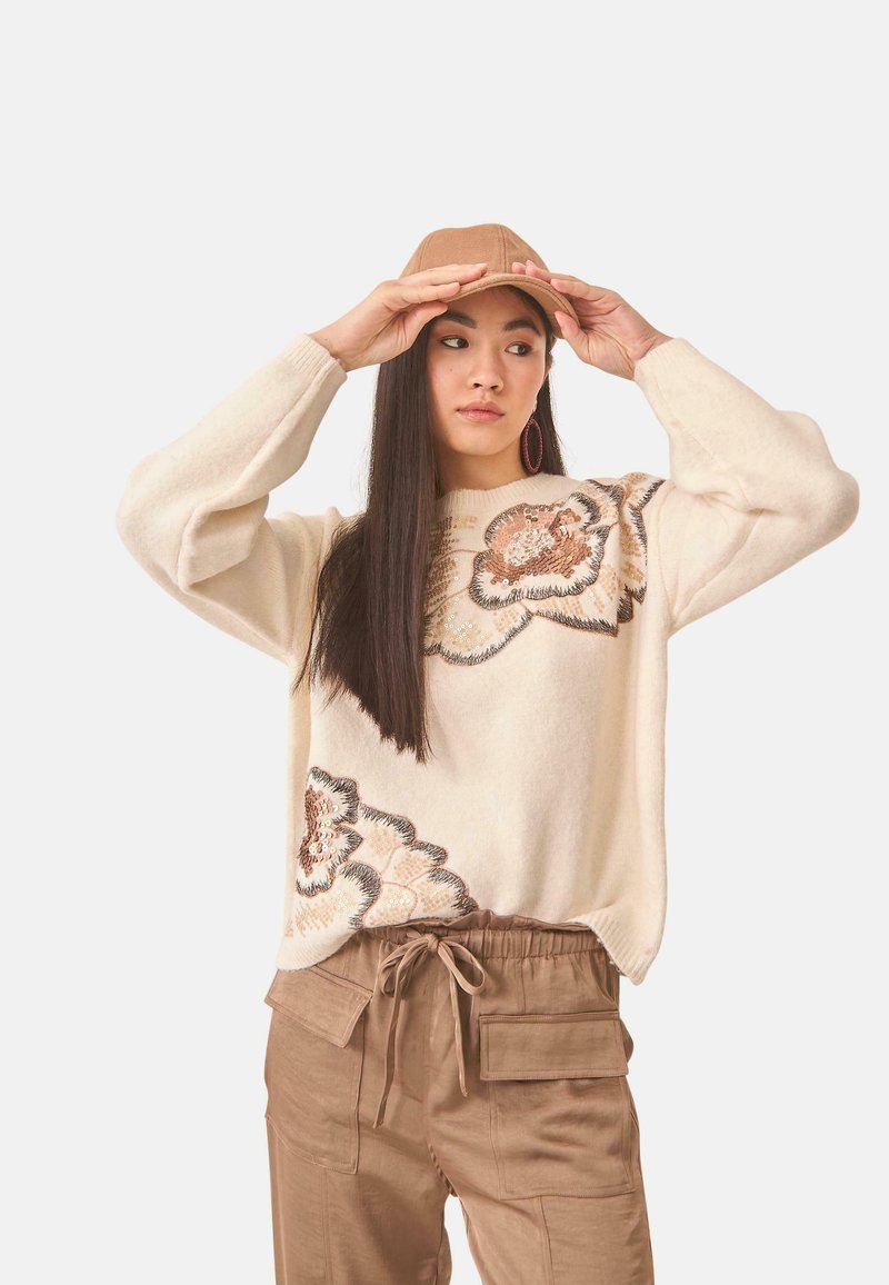 Suéter crema con grandes diseños florales en marrón y negro, combinado con pantalones beige. El modelo lleva una gorra a juego en tono marrón.