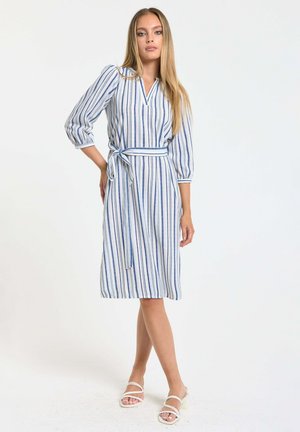 Woman standing wearing a knee-length white dress with vertical blue stripes, three-quarter sleeves, a tied waist belt, and white heeled sandals.