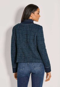 Frau in einem strukturierten marineblauen Blazer mit dunklen, umgeschlagenen Ärmeln und blauen Jeans, seitlich vor einem schlichten Hintergrund stehend.