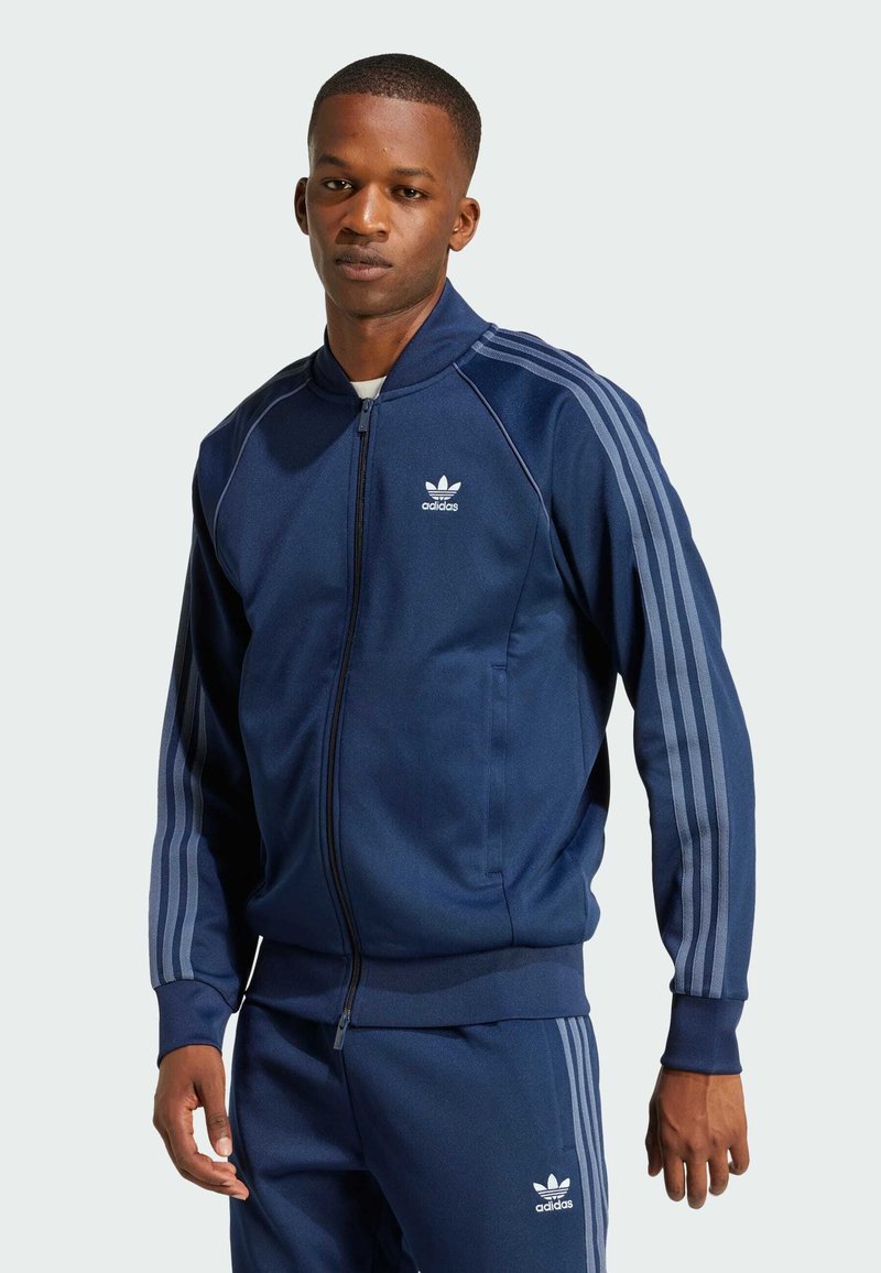 Navy blue track jacket made of smooth fabric, featuring a full zip, ribbed cuffs, and three light gray stripes down each sleeve.