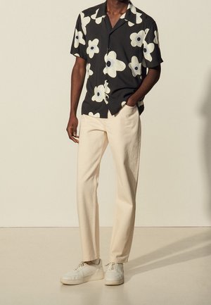 Mężczyzna w czarnej koszulce z krótkim rękawem ozdobionej dużymi białymi kwiatami, beżowych spodniach i białych trampkach, stoi z jedną ręką w kieszeni.